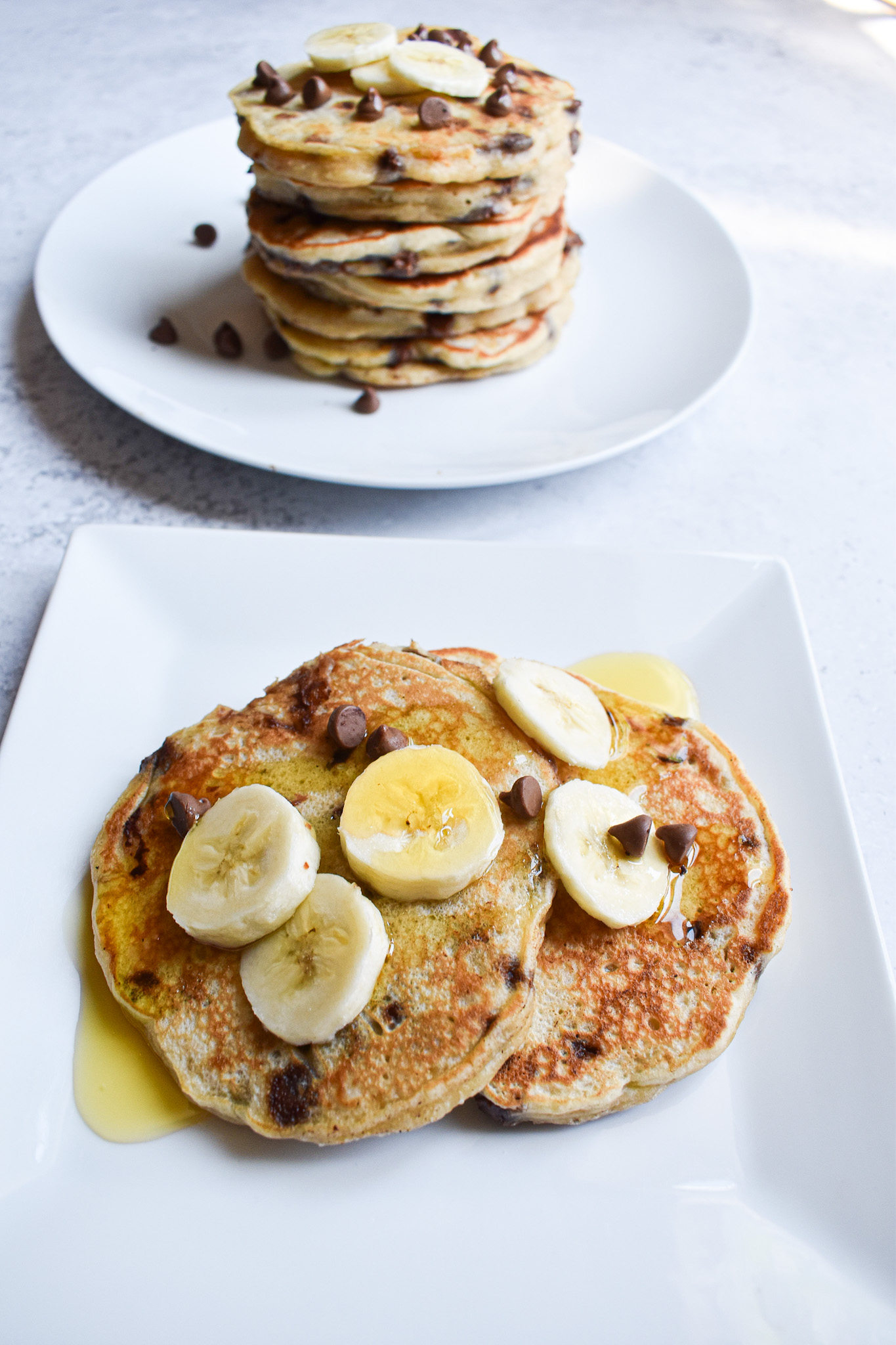 A white plate with 2 pancakes with syrup and chopped bananas. A stack of pancakes behind the  front plate.