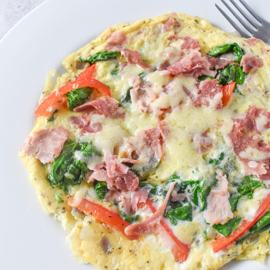 Omelette on plate topped with spinach, red peppers, ham and cheese. A fork is next to the omelette.