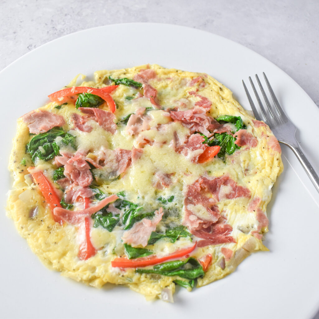 Omelette on a white plate topped with ham, spinach and red peppers and a silver fork next to the omelette.