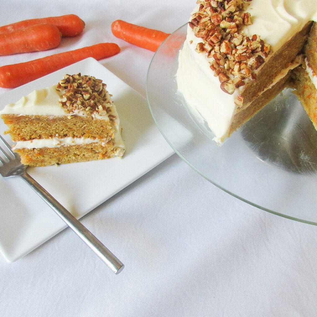 A slice of carrot cake on plate with a silver fork and to the right a cake on a turntable.